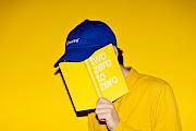 A man wearing a yellow shirt and blue cap covering his face with a yellow planner in front of a yellow background