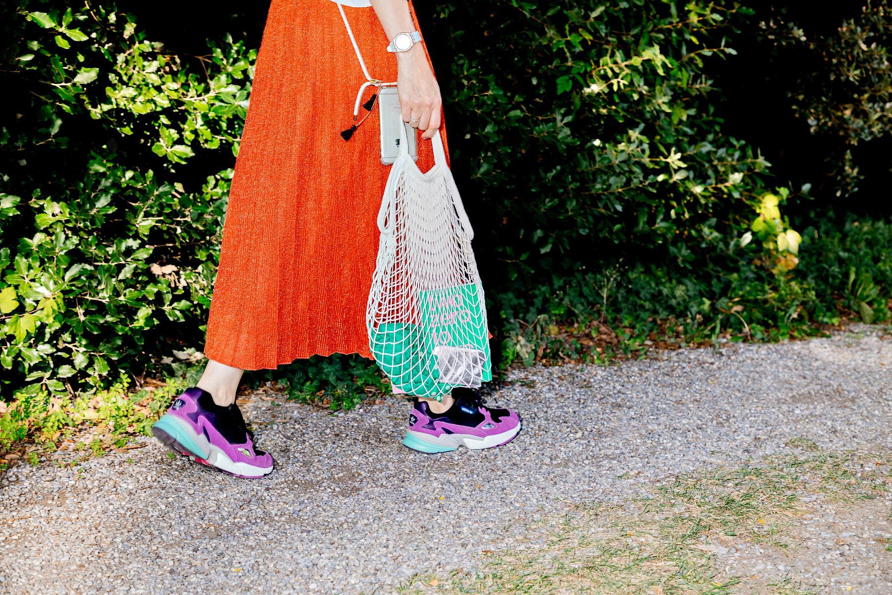 A fashionable woman walking on a path in a park holding a net shopping bag