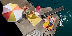 Two people lounging on deck chairs on a pier by lake Zurich. There is a colourful SULA x Micasa umbrella providing shade, and SULA xMicasa towels spread out on the pier.
