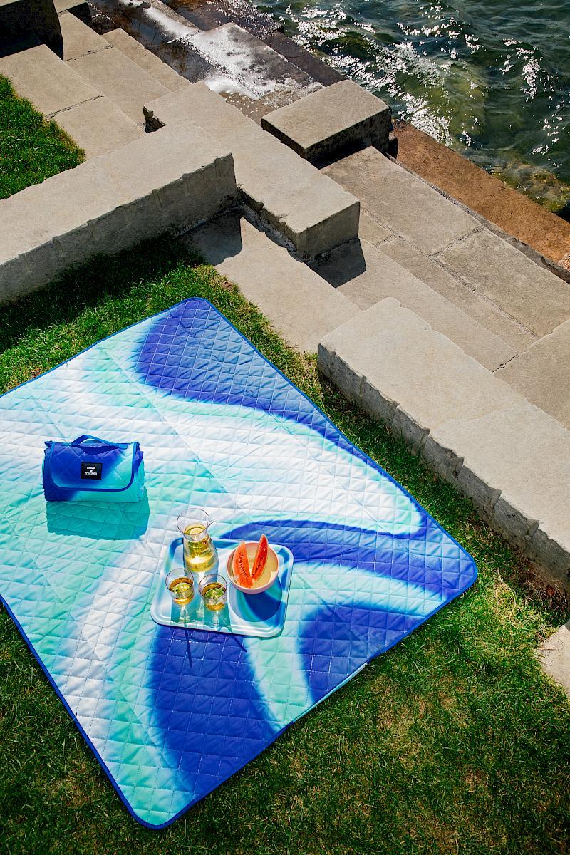 A picnic scene set on a grassy area near a Lake Zurich. A vibrant blue and green SULA x Micasa picnic blanket is spread out on the grass. On the blanket, there are two glasses of drink, slices of watermelon, and a tray. Stone steps lead down to the water