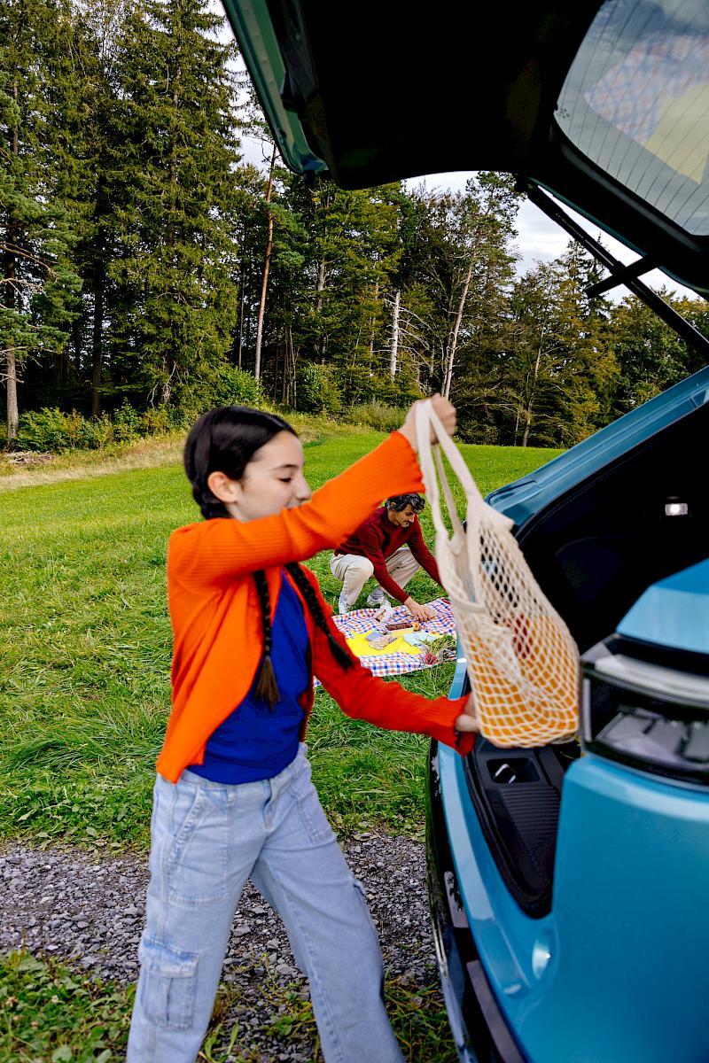 Child placing tote bag into the trunk of a blue electric SUV while an adult packs up a picnic in the background