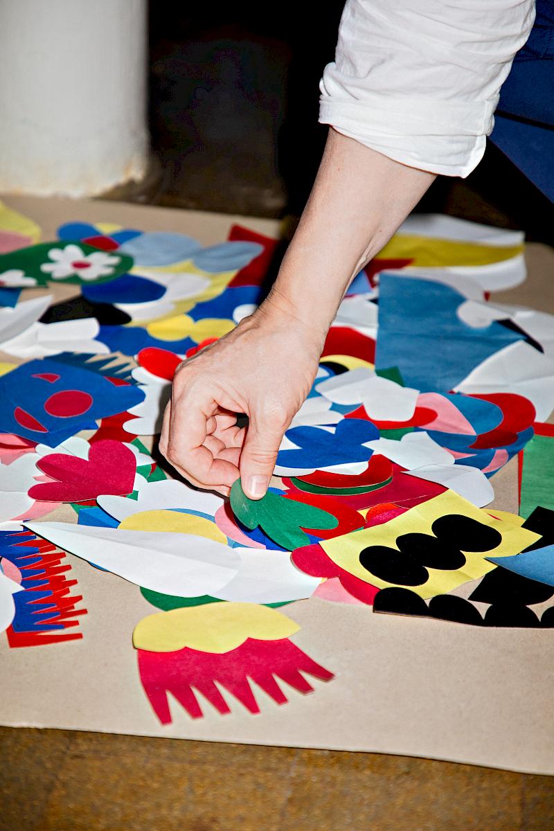 Close-up of a hand arranging colourful paper cut-out shapes on a large worktable inside an artist's studio